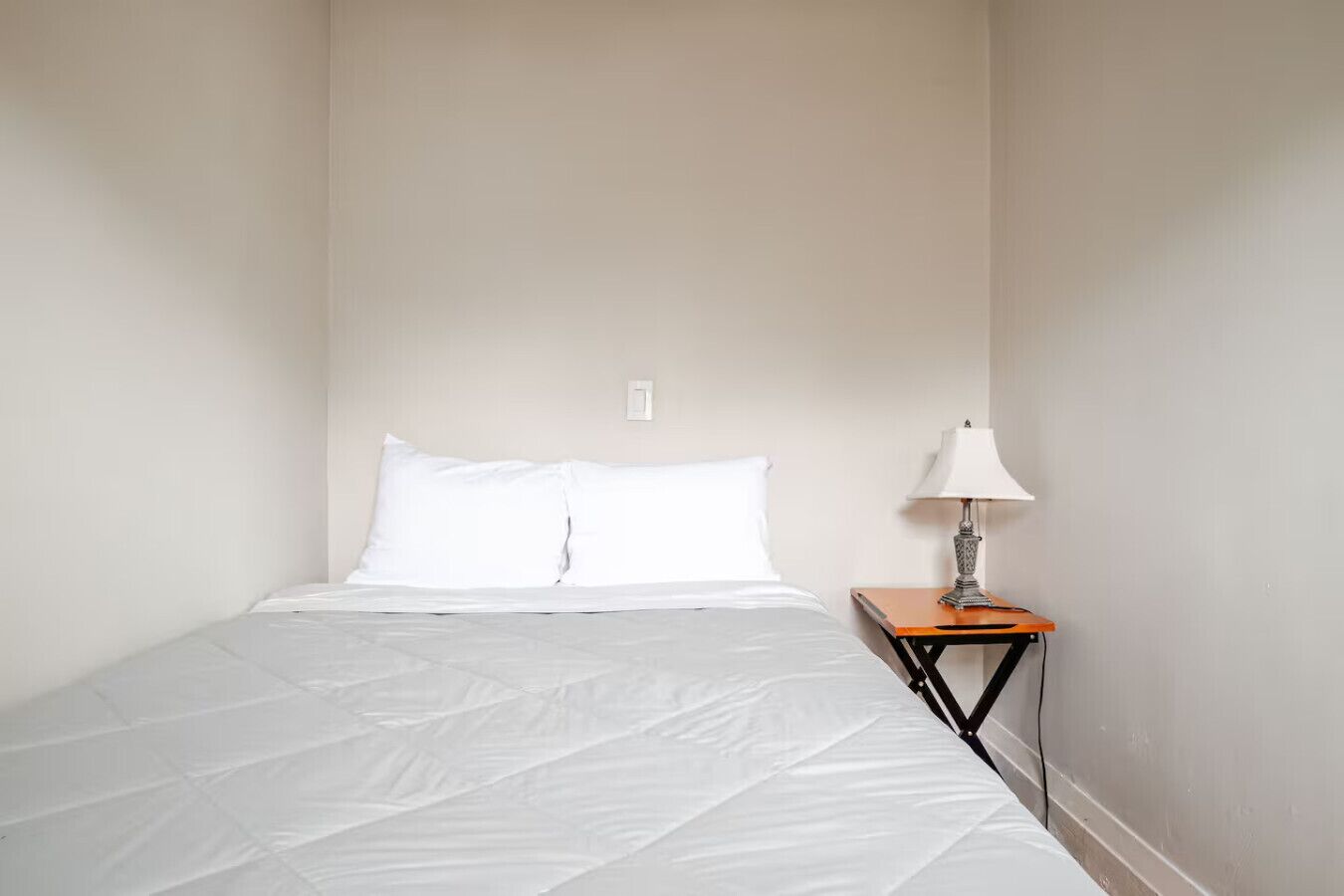 Una cama individual con ropa blanca en una habitación de paredes sencillas y una mesita de noche de madera pequeña con una lámpara.