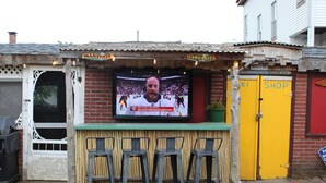 Una terrassa de bar amb una pantalla de televisió, un taulell de fusta i tres tamborets.