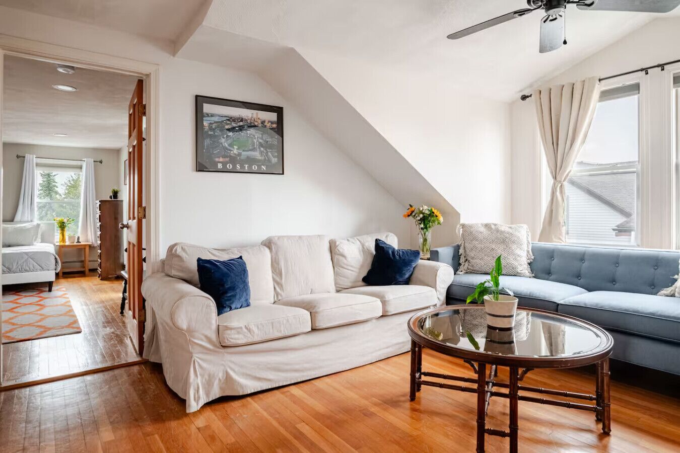 Een moderne woonkamer met een witte bank, een blauwe bank, een glazen salontafel en een schilderij van Boston aan de muur.
