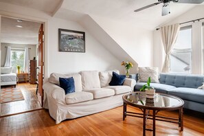 Un salon moderne avec un canapé blanc, un canapé bleu, une table basse en verre et un tableau représentant Boston accroché au mur.