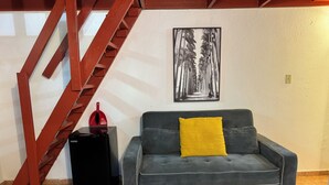 A grey sofa with a yellow pillow, a red fire extinguisher, a black suitcase, and a wooden staircase.
