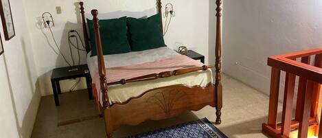 A four-poster bed with a wooden frame, a green pillow, and a white bedspread. There is a small bedside table with a lamp, a blue patterned rug, and a wooden railing.