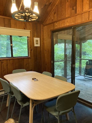 Ein Zimmer mit Holzwänden, einem großen Tisch und Stühlen, einer Kristallleuchter und Blick ins Freie durch eine Glastür.