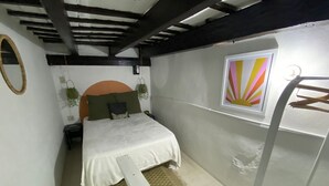 A bedroom with a bed, a nightstand, a wall-mounted shelf, a hanging plant, and a window with a sunburst design.