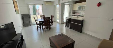 A modern living room with a TV, a wooden coffee table, and a sofa. There is a dining area with a table and chairs, and a kitchen with a fridge and a sink.