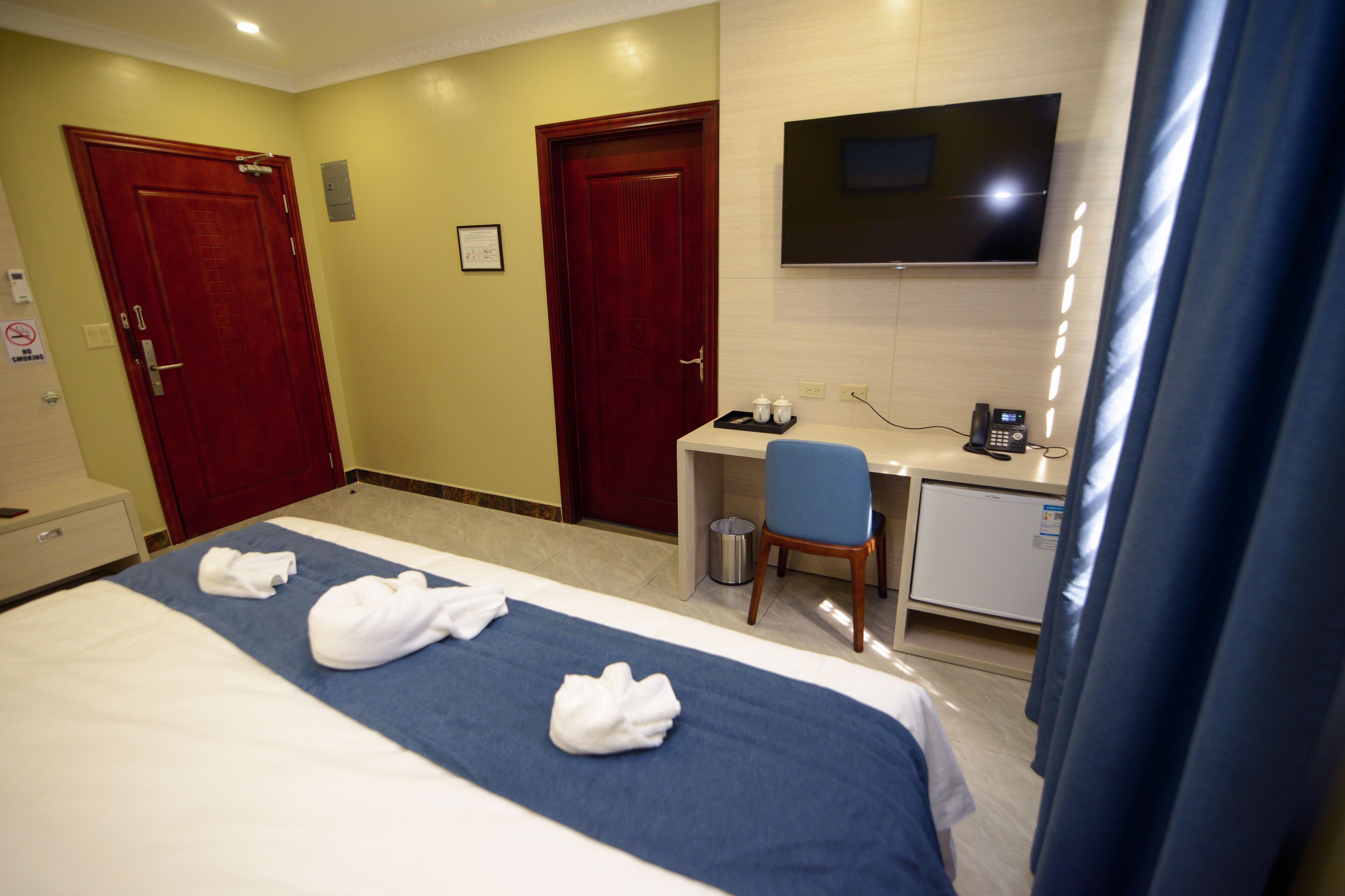 A hotel room with a bed, two doors, a desk with a chair, a TV, and a trash can.