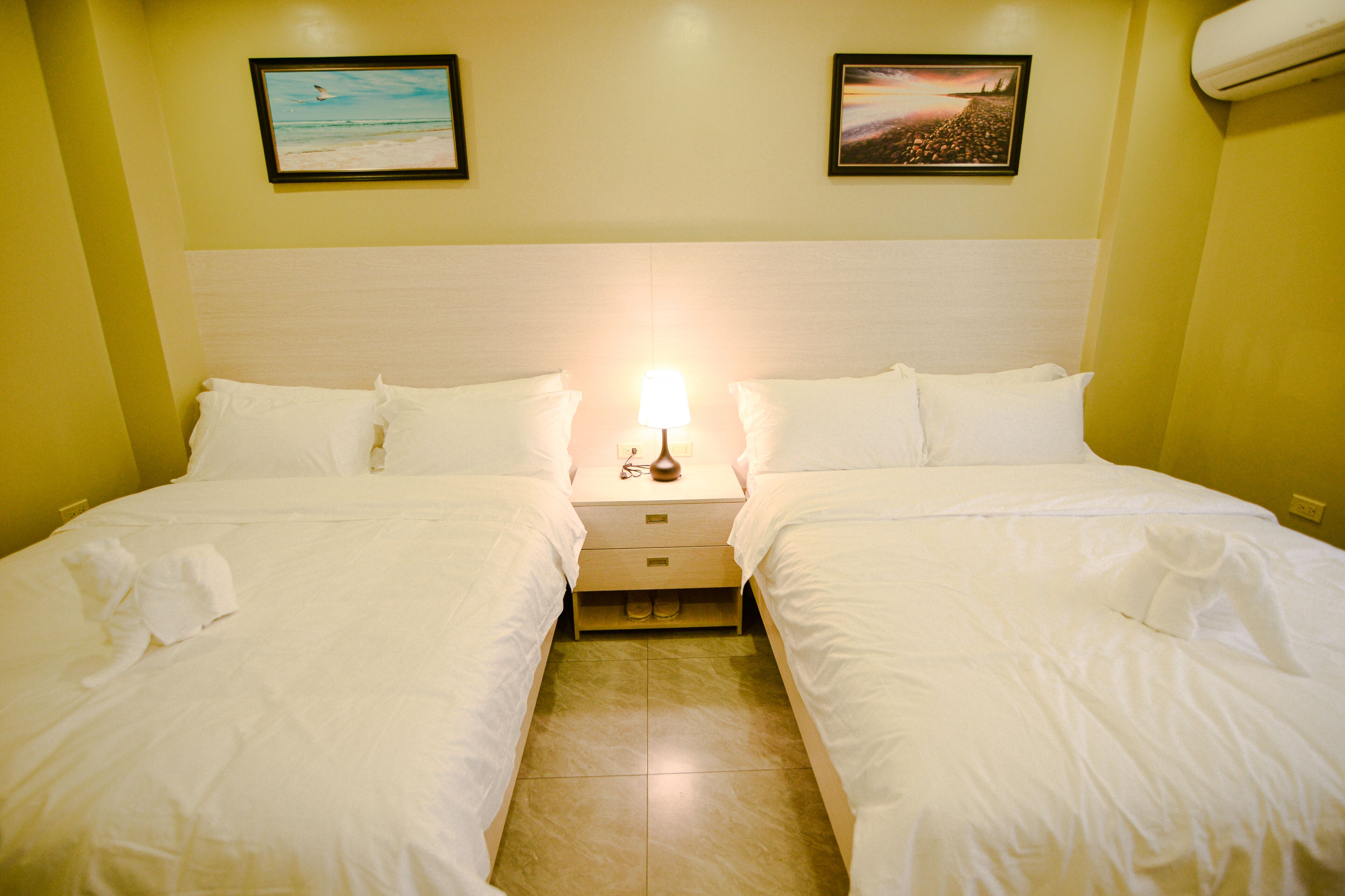 A hotel room with two beds, a nightstand with a lamp, and two framed pictures on the wall.