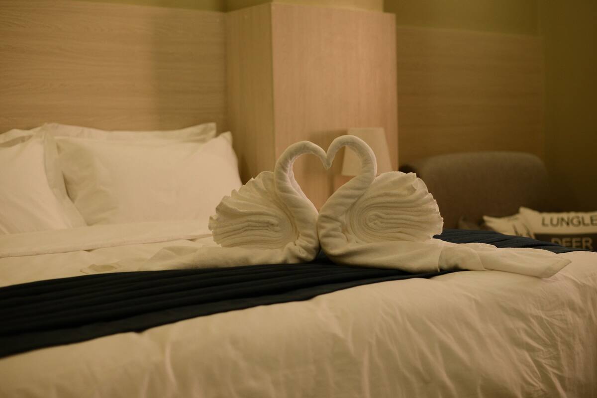 A hotel room with a neatly made bed, a heart-shaped towel arrangement, and a pillow with text.