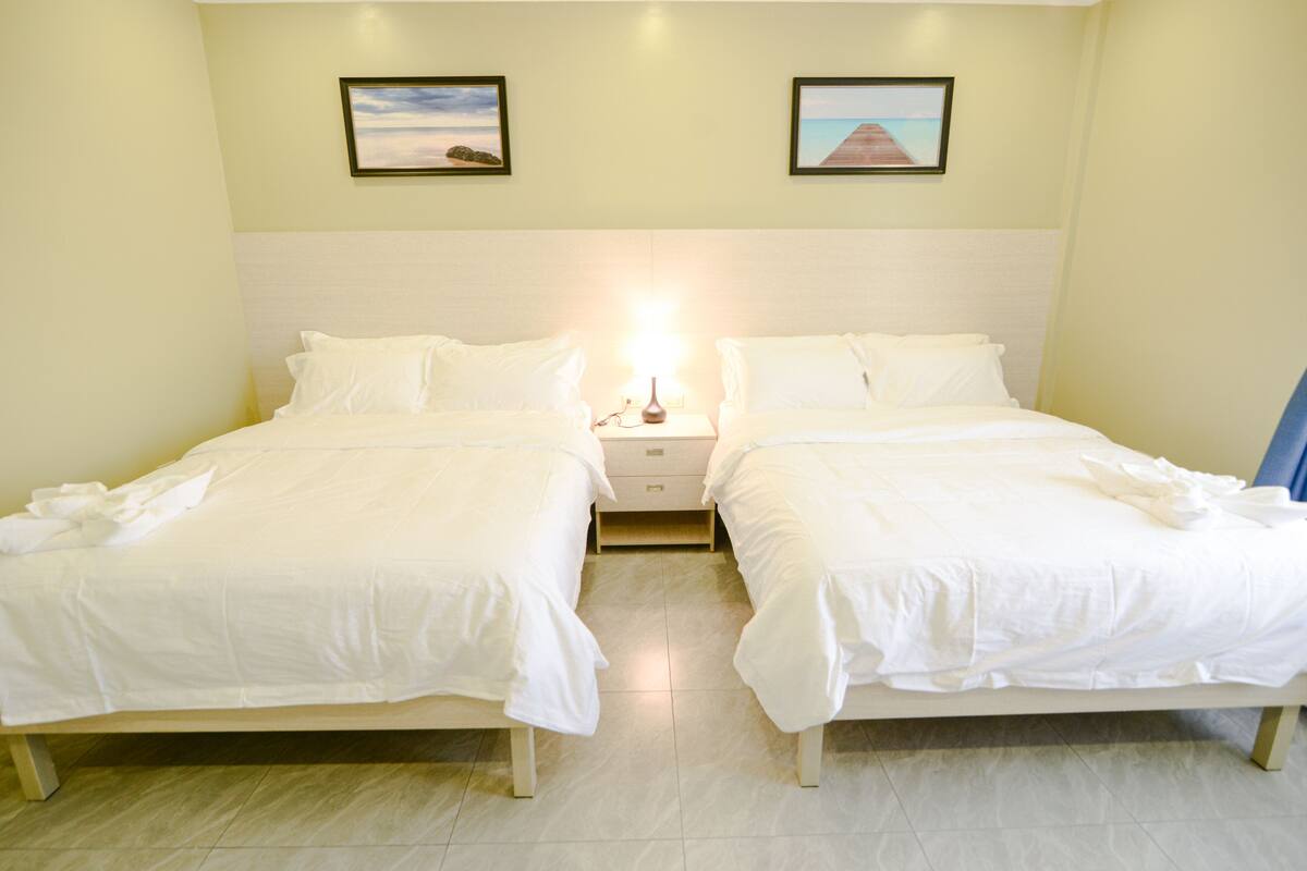 A hotel room with two single beds, a nightstand with a lamp, and two framed pictures on the wall.