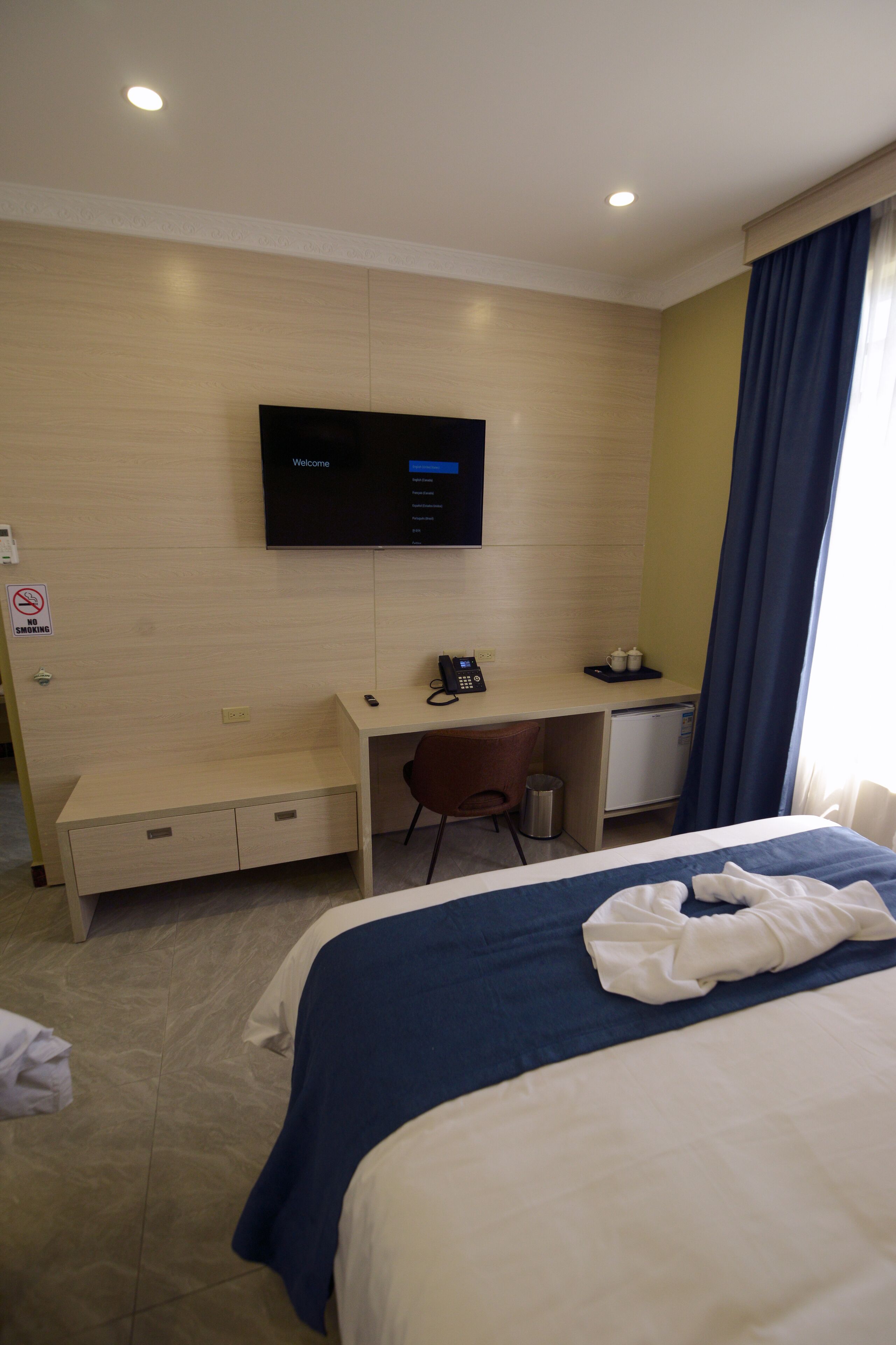A hotel room with a bed, a desk, a television, a mini-fridge, and a no-smoking sign.