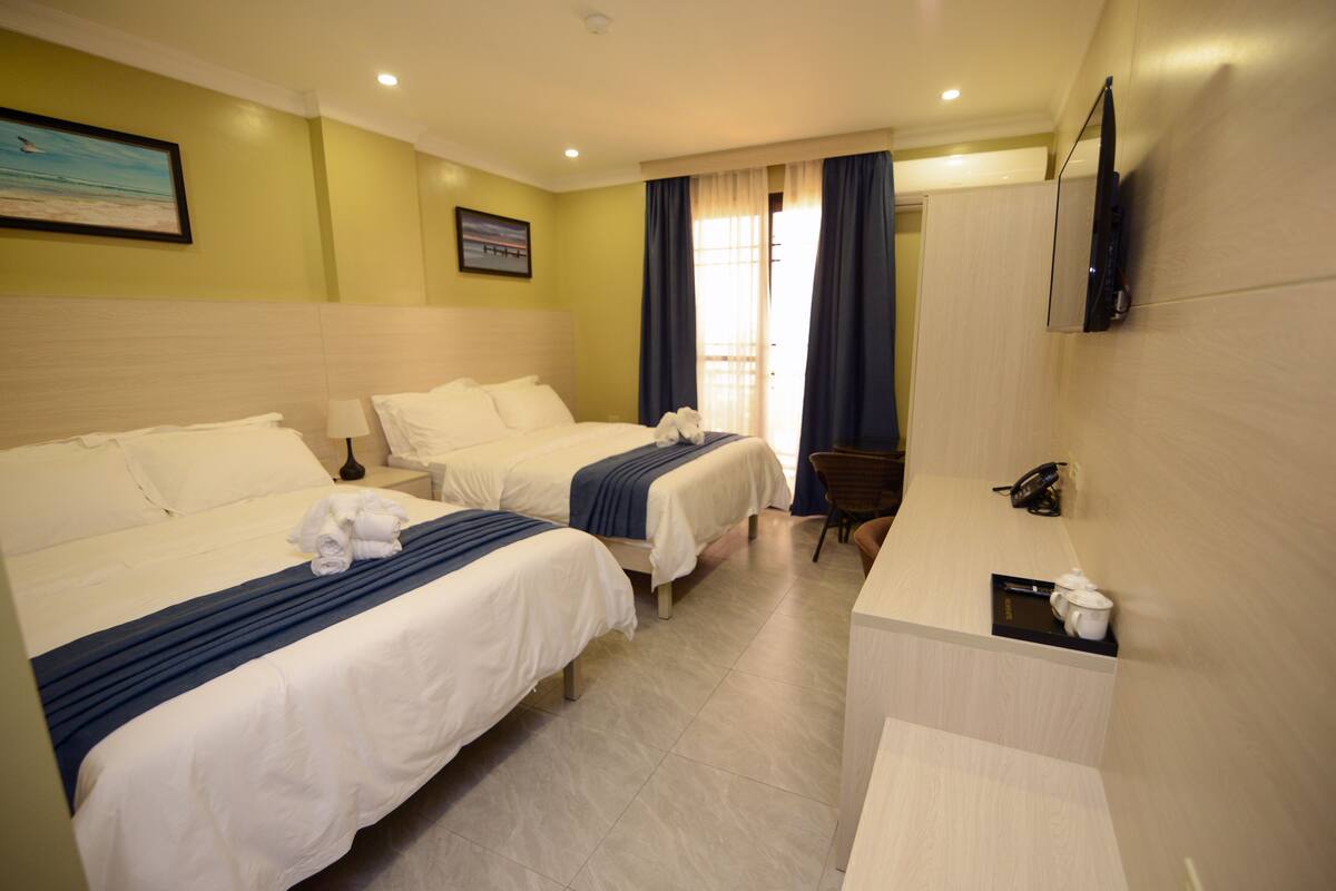 A hotel room with two beds, a desk, a chair, a TV, and a window with curtains.