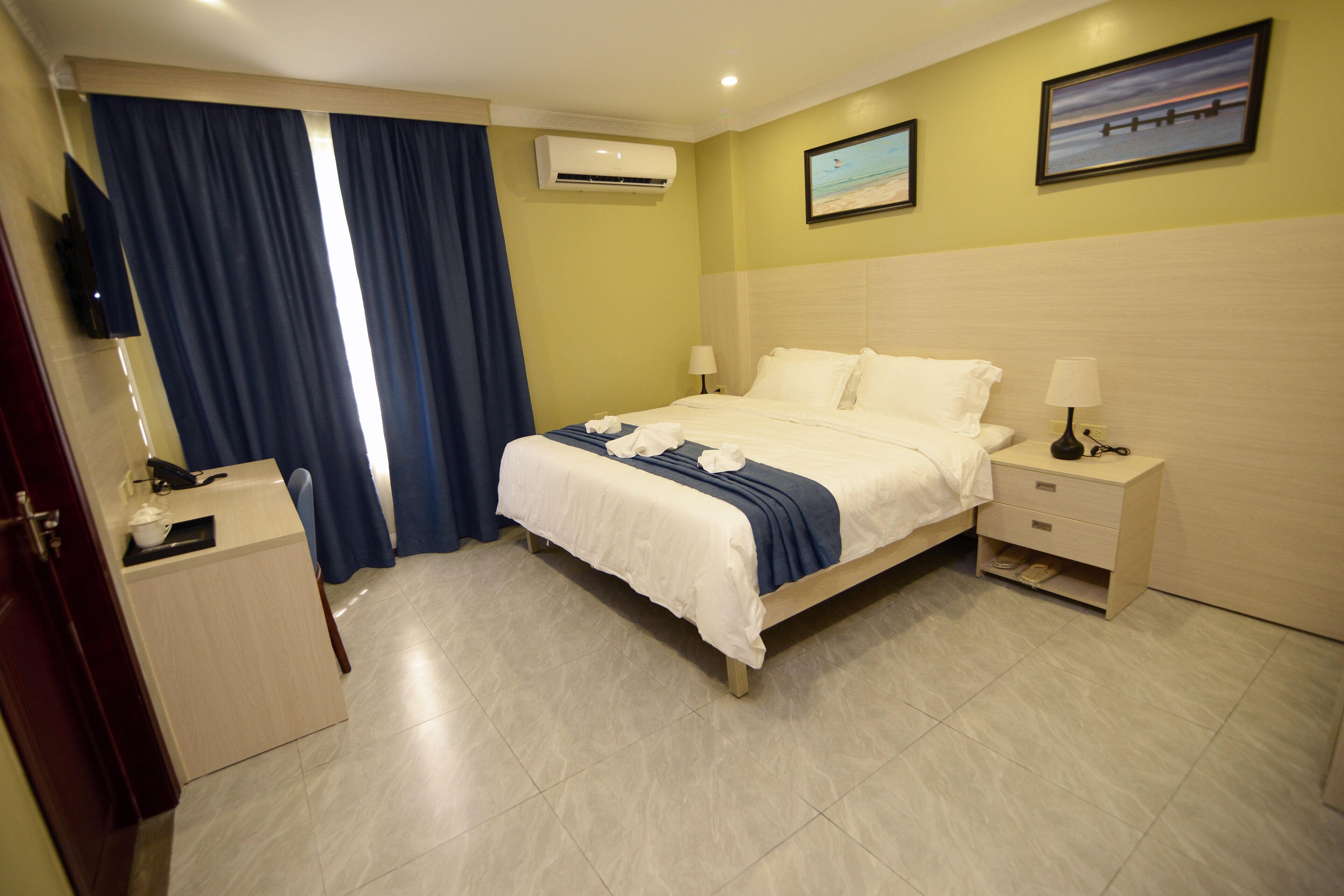 Una habitación de hotel con cama, mesitas de noche, un escritorio, un equipo de aire acondicionado y un cuadro en la pared.