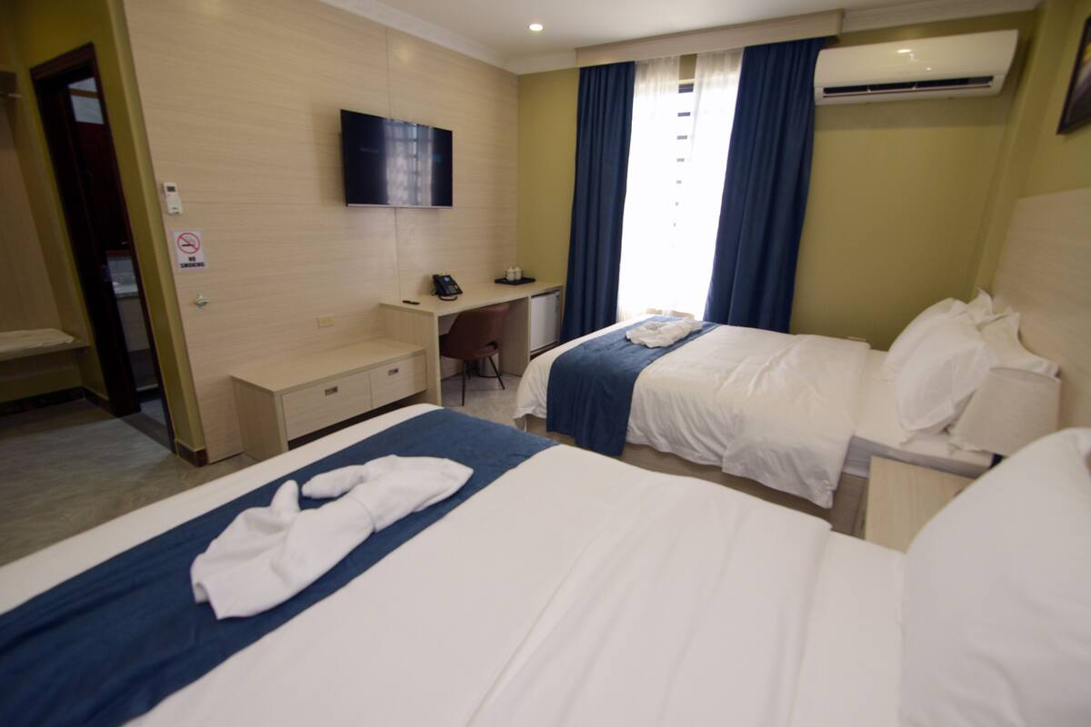A hotel room with two beds, a desk, a chair, a television, and an air conditioning unit.