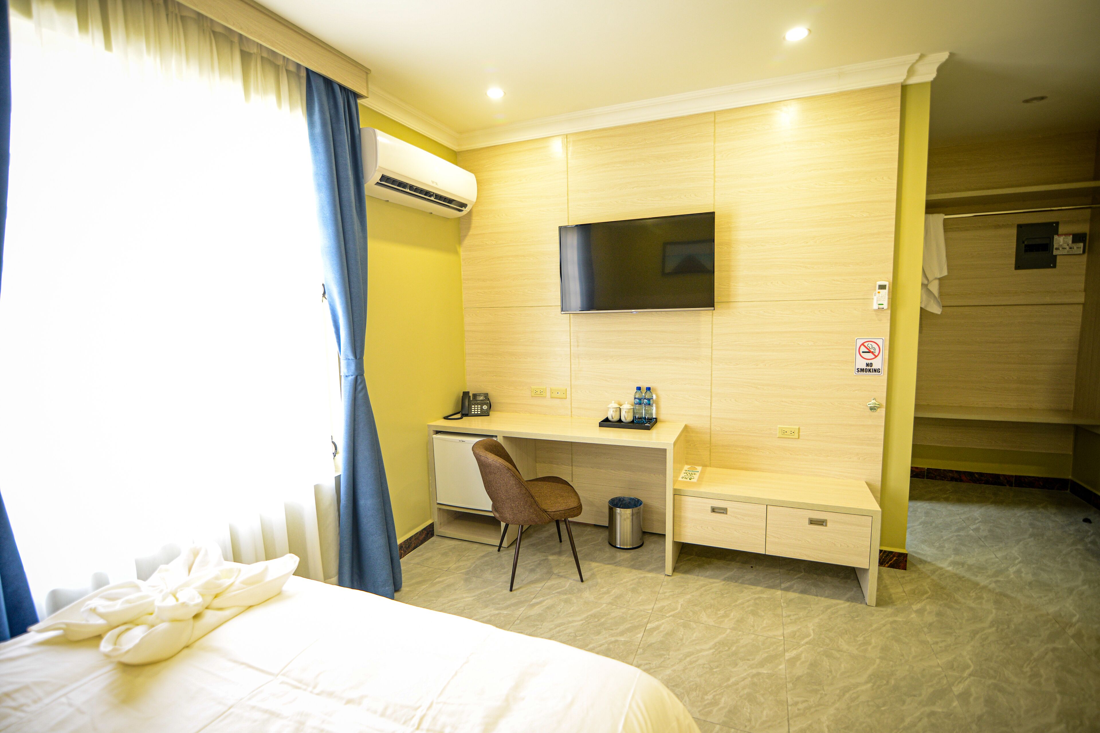Een hotelkamer met een bed, een bureau met stoel, een aan de muur bevestigde televisie en een airconditioning.