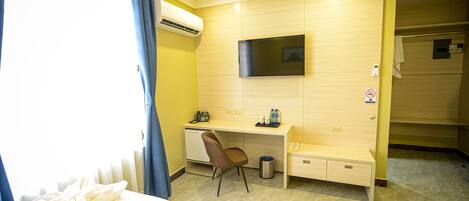 Een hotelkamer met een bed, een bureau met stoel, een aan de muur bevestigde televisie en een airconditioning.