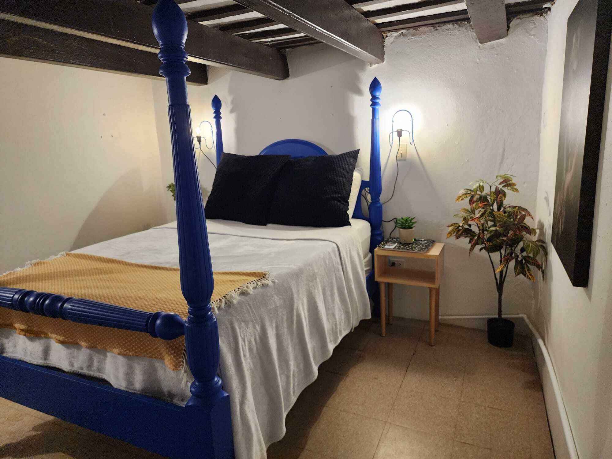 A bedroom with a four-poster bed, a bedside table with a lamp, and a potted plant.