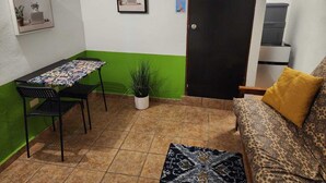 Un petit espace de vie compact comprenant une table à manger, deux chaises, un canapé avec un coussin jaune, une plante en pot et un tableau encadré accroché au mur.