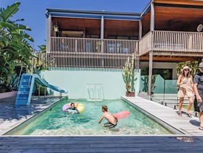 Ein Schwimmbecken mit einer Rutsche, umgeben von einer Holzterrasse und einem Gebäude mit einem Balkon.
