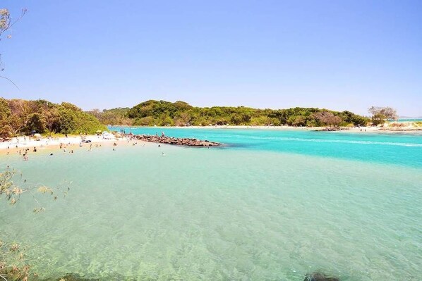 Ein Strand mit klarem türkisblauem Wasser, sandigem Ufer und üppigen grünen Hügeln im Hintergrund.