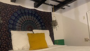 Una cama con ropa de cama blanca y una almohada color mostaza, un tapiz con diseño de pavo real en la pared, y una planta colgante.