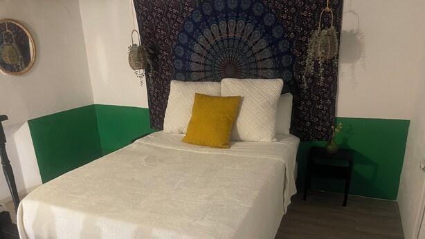 Una cama con una colcha blanca y un almohadón amarillo, una pared verde y blanca, un tapiz y una pequeña mesa con un florero.