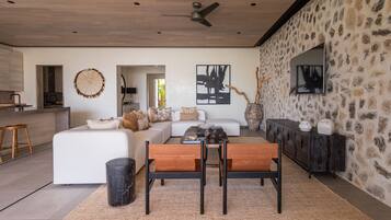 A modern living room with a white sofa, a dining table with chairs, a black trash bin, and a stone wall with artwork.