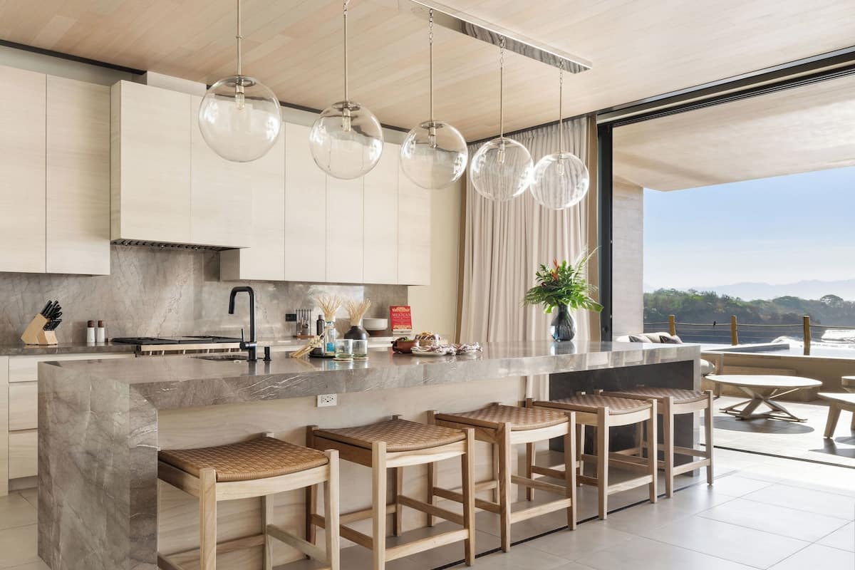 Una cocina moderna con una isla de mármol, taburetes de bar, luces colgantes y vistas al océano.