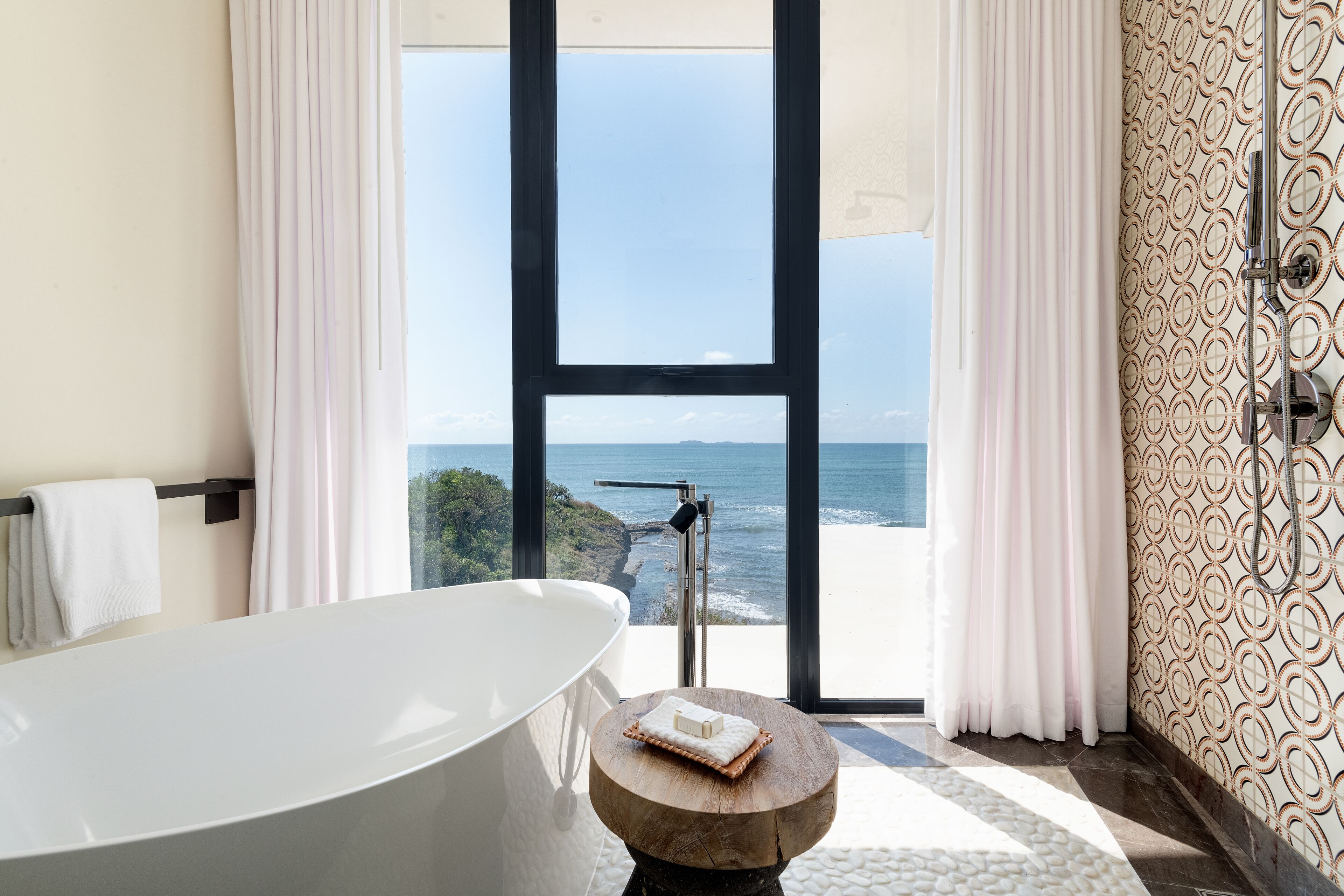 Un baño moderno con una bañera independiente, vistas al océano y un papel tapiz estampado.