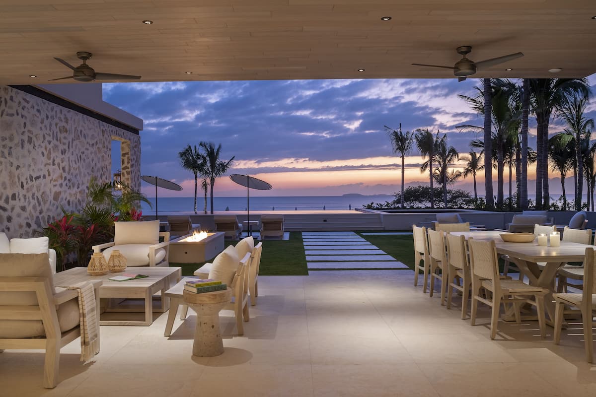 Un patio exterior con mobiliario, una fogata y vistas al océano al atardecer.