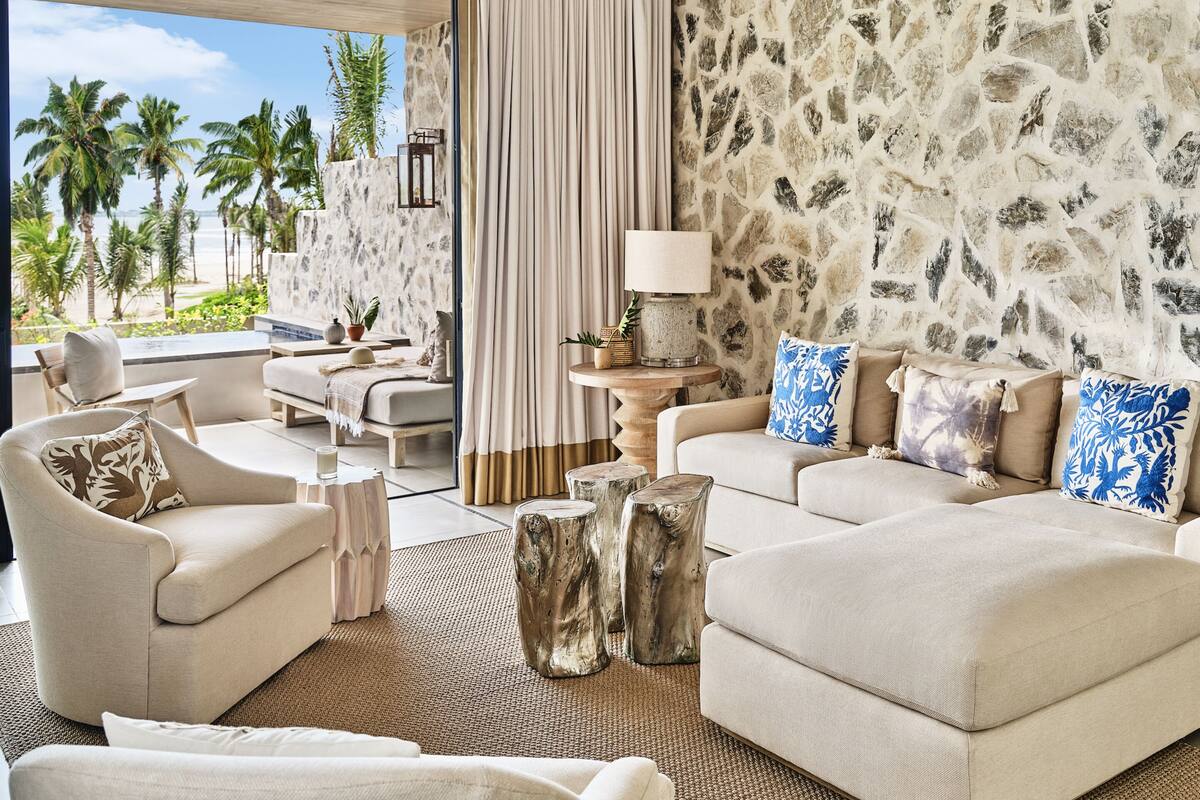 Una sala de estar moderna con pared de piedra, un conjunto de sofás, una mesa de centro y vistas a palmeras y al océano.