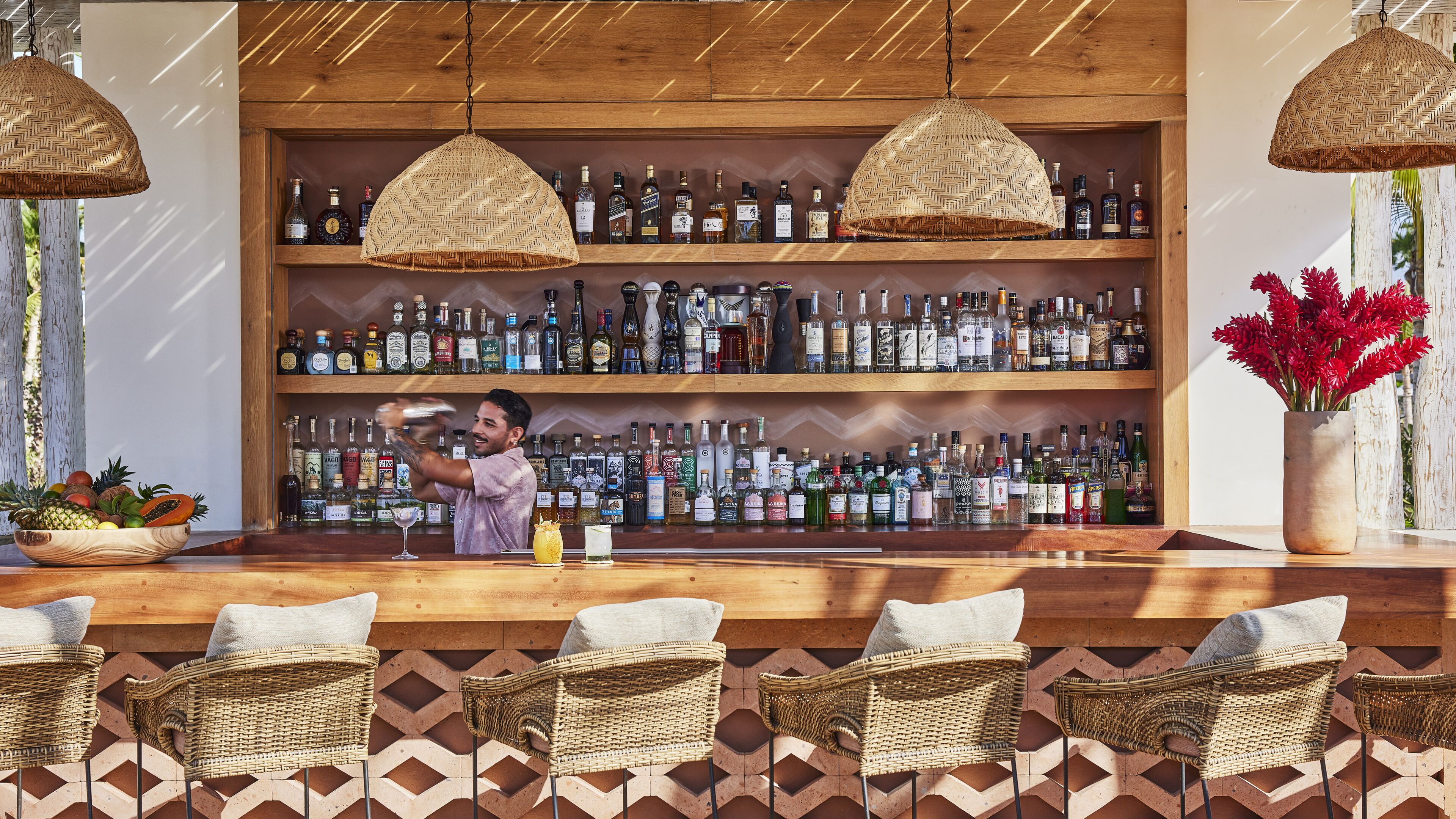 Een bar met rieten stoelen, een bartender die drankjes maakt en schappen vol drankflessen.