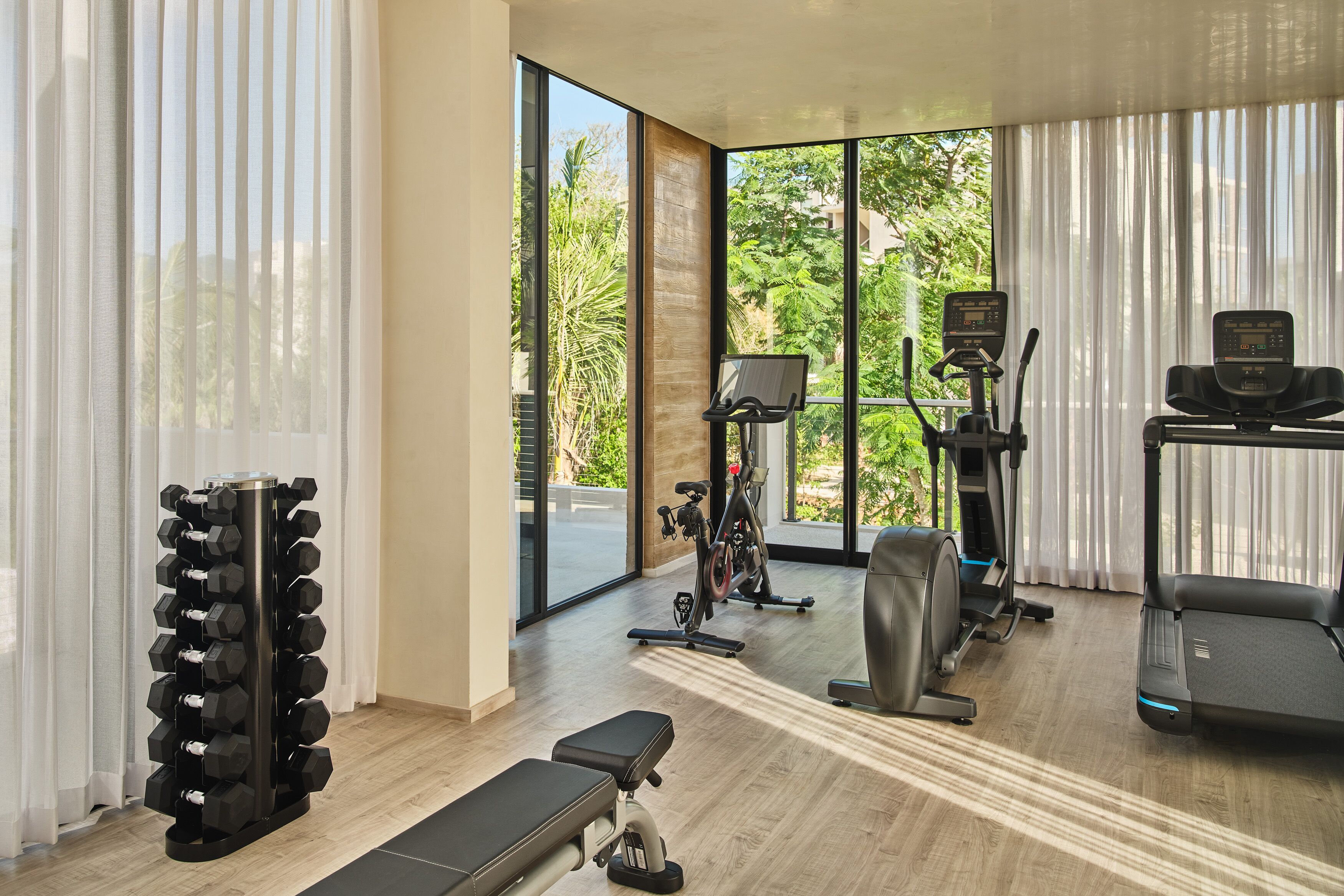 A modern gym with exercise equipment, large windows, and a view of greenery.