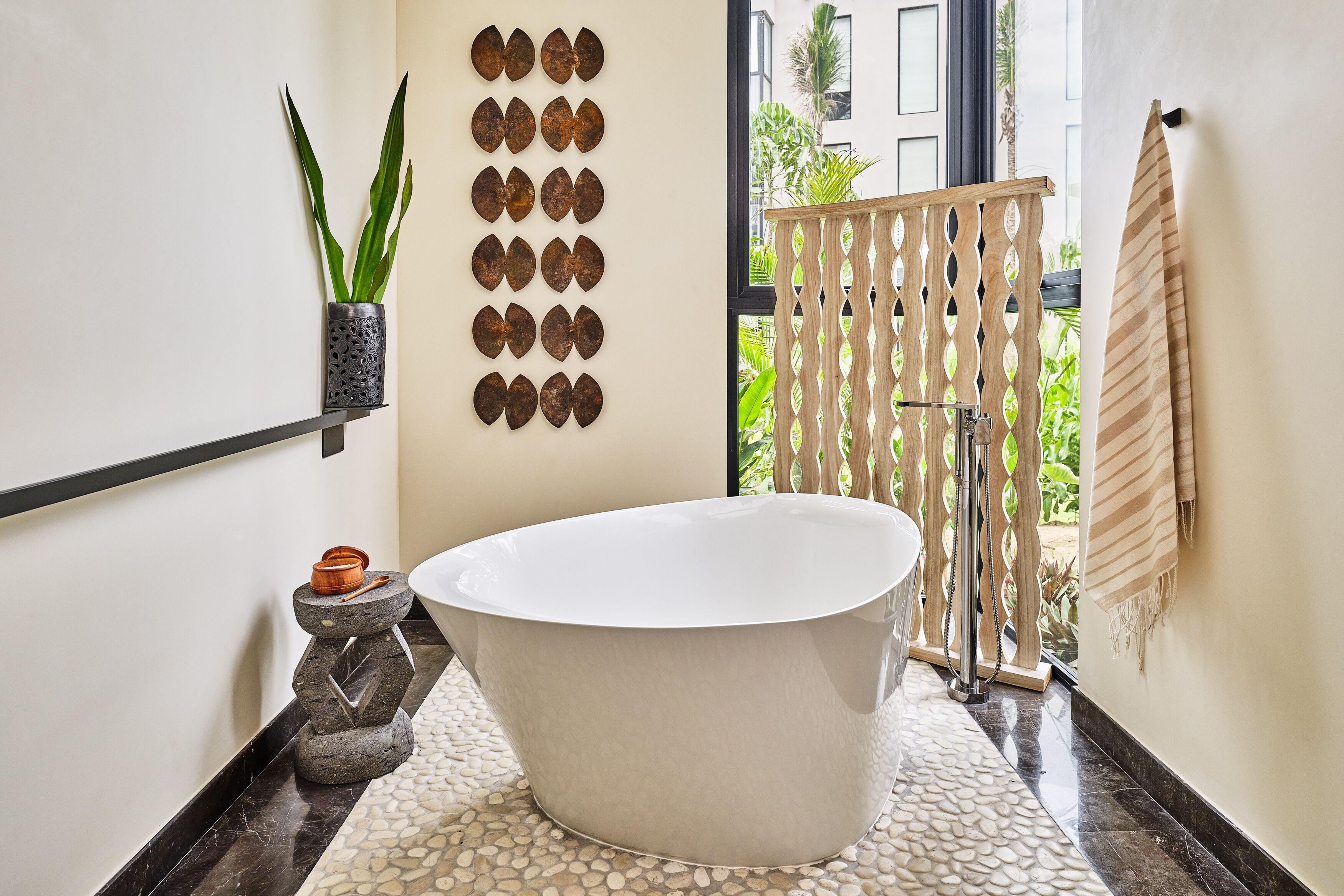 A modern bathroom with a freestanding bathtub, a potted plant, decorative wall art, and a view of the outdoors.