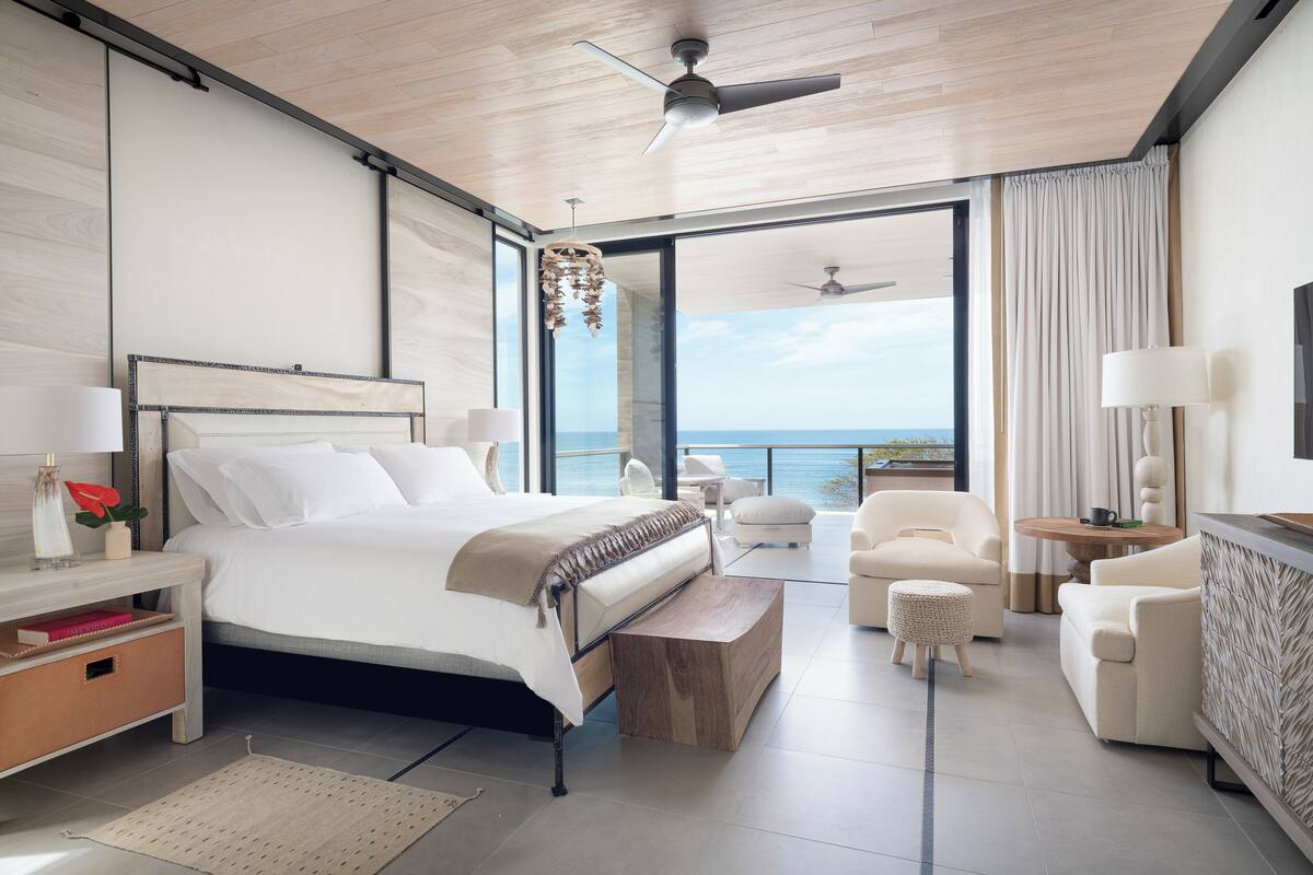 Un dormitorio moderno con una cama grande, ventilador de techo, vista al océano y balcón con mobiliario.