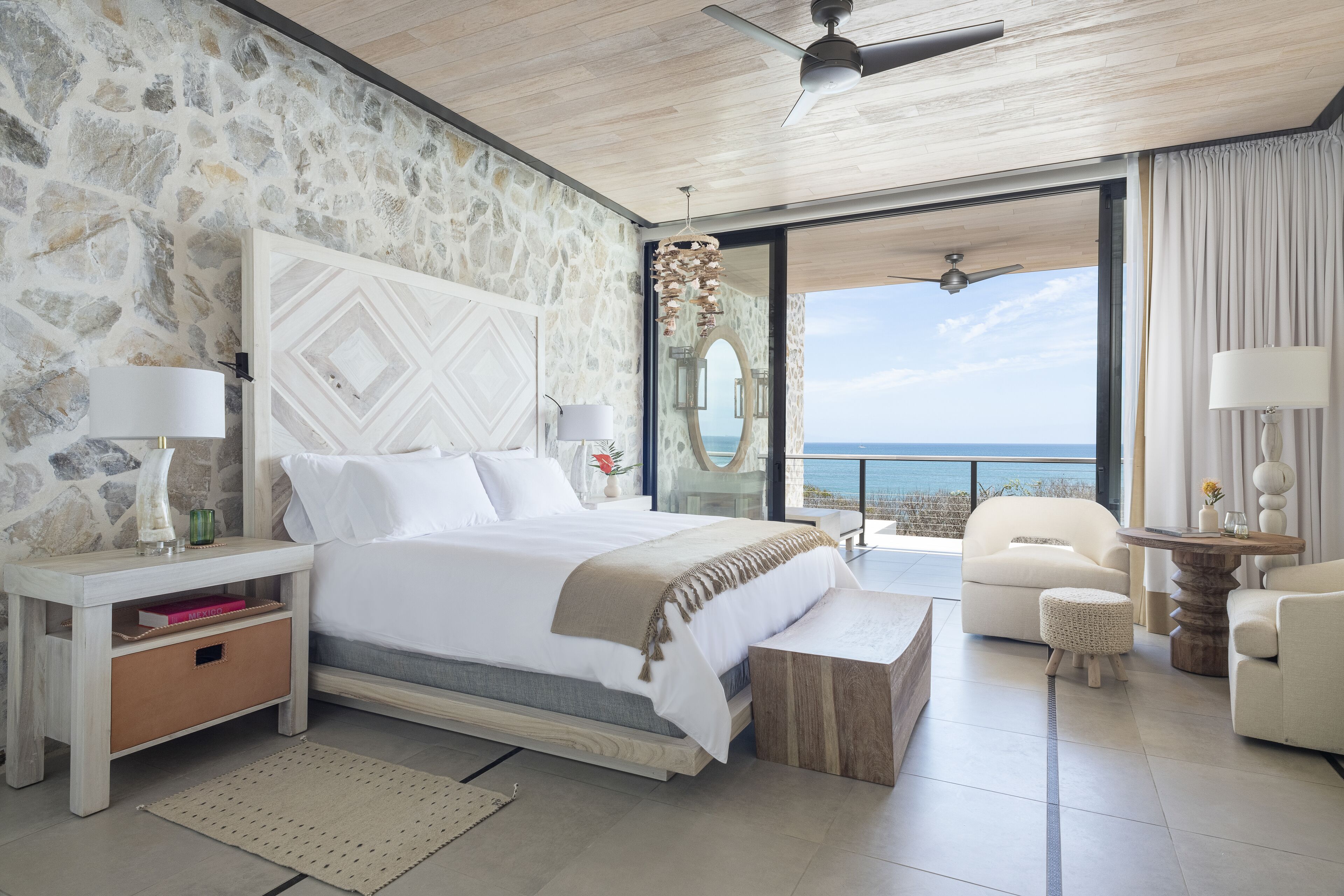 Un dormitorio con pared de piedra, una cama con ropa de cama blanca, una mesita de noche y lámparas, vista al mar, y un ventilador de techo.