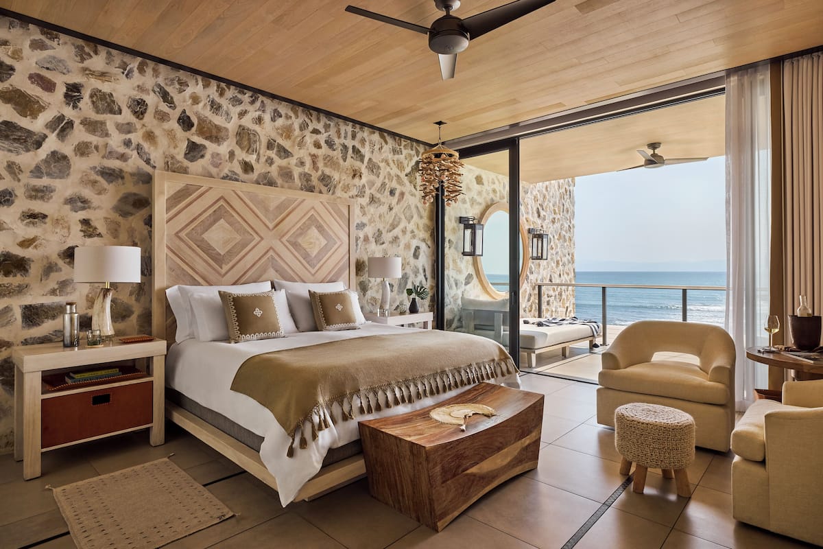 Un dormitorio con pared de piedra, cama con cabecera estampada, mesa de centro de madera, una silla y vista al océano.