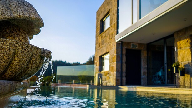 A stone fountain with water spouting from its center, a swimming pool in the foreground, and a modern stone building with large windows and a flat roof in the background.