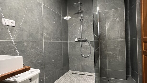 Un baño moderno con azulejos grises, cabina de ducha de vidrio, lavamanos blanco y estante de madera.