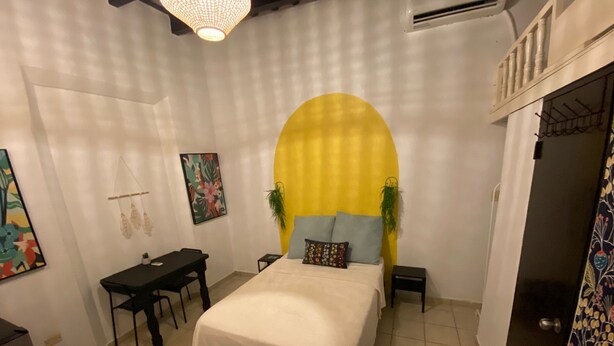 A bedroom with a bed, a desk, a chair, a hanging plant, and a door with a patterned curtain.