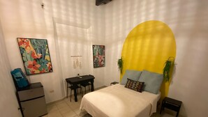 A bedroom with a bed, a desk, and two chairs. There is a large yellow wall and a white door.
