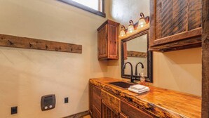A bathroom with a wooden vanity, a sink, a mirror with lights, and a towel rack. - Mountain Lion Den at Snowcat Cabins (Hot Tub!) (Coram)