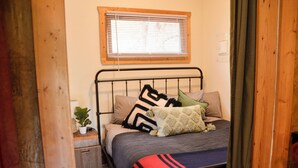 A bedroom with a bed, a bedside table with a plant, and a window with blinds. - Marmot Mini Cabin (Unit 7) at The Woolly Bugger Inn (Estes Park)