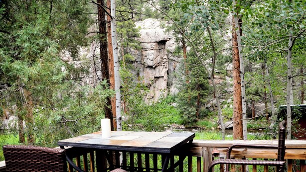 An outdoor patio with a table and chairs, surrounded by trees and a rocky cliff. - Marmot Mini Cabin (Unit 7) at The Woolly Bugger Inn (Estes Park)