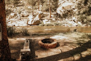 Un terrain de camping avec un foyer, un banc et une rivière en arrière-plan.