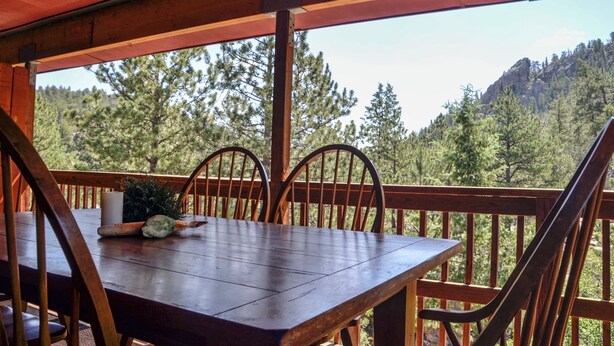 A wooden deck with a table and chairs, overlooking a forested mountain area.