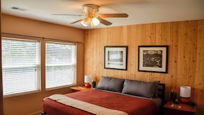 A bedroom with a bed, two bedside tables, a ceiling fan, and two windows with blinds.