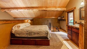 Ein Schlafzimmer mit Holzbalkendecke, einem Bett mit gemustertem Überwurf, einem Nachttisch, einer Kommode und einem Spiegel.