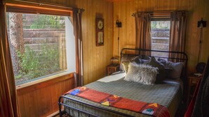 A wooden-paneled room with a bed, a window with curtains, a wall clock, and a small table with a lamp. - Canyon Cabin (Unit 17) at The Woolly Bugger Inn (Estes Park)