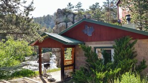 A rustic cabin with a Texas state flag on the front, a wooden deck, and a view of a river and forested mountains. - Canyon Cabin (Unit 17) at The Woolly Bugger Inn (Estes Park)