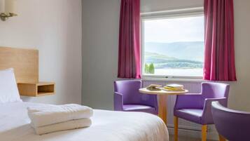 Una habitación de hotel con una cama, dos sillones morados, una mesita y un ventanal con cortinas.